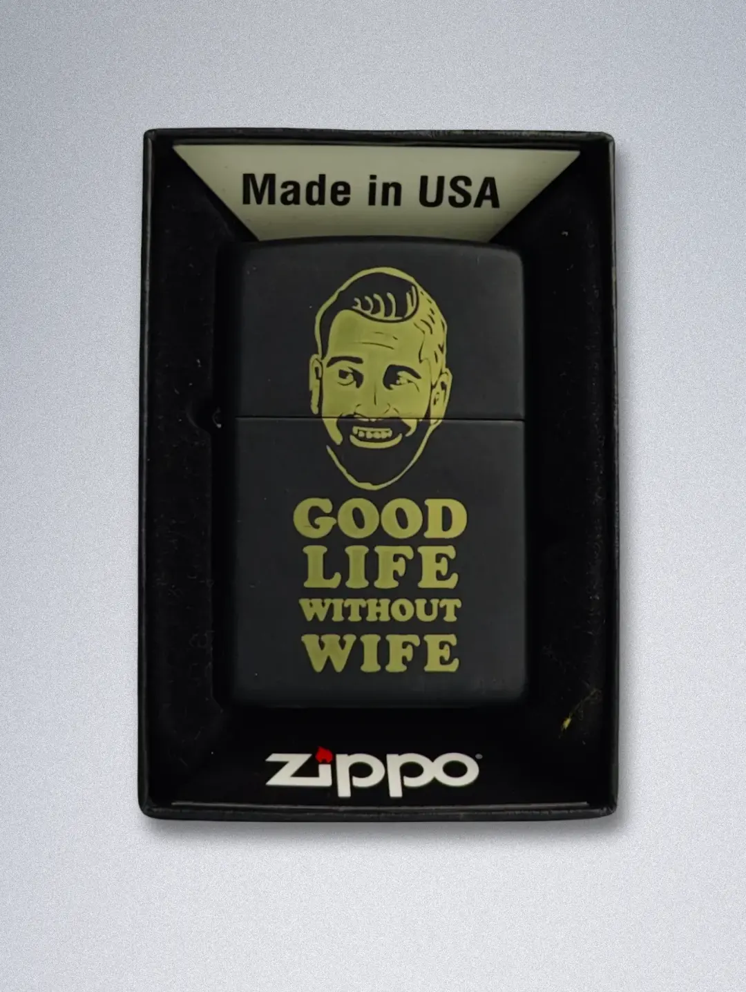75280 (2) A matte black Zippo lighter with a gold graphic of a smiling, bearded man and the text "GOOD LIFE WITHOUT WIFE".